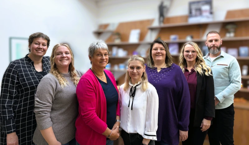 Ein Team von sieben Mitarbeitenden steht lächelnd in einem modernen Büro mit Bücherregalen.